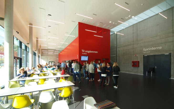 Cafenea spațioasă cu decor norvegian modern, cu oameni care stau la coadă lângă un perete roșu etichetat "Til ungdommen" și alții așezați la mese lângă ferestre mari.