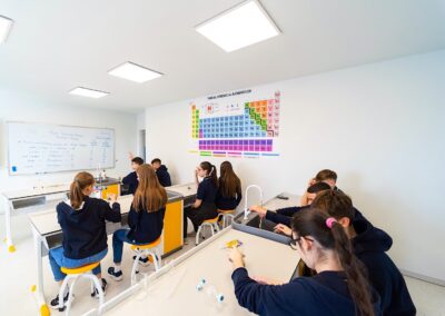 Elevii de la o scoala privata din sectorul 3 stau la mesele de laborator dintr-o clasă de științe, lucrând la activități cu echipamente de laborator; un tabel periodic atârnă pe perete, iar pe tablă sunt scrise notițe.