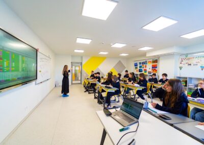 Un profesor stă lângă o tablă digitală, prezentând elevilor cu laptopuri într-o sală de clasă modernă și luminoasă din scoala privata, prezentând mediul de învățare captivant al unei scoala privata din sectorul 3.