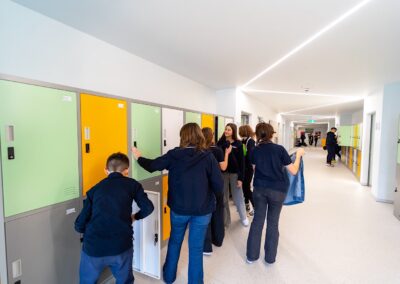 Elevii interacționează în apropierea dulapurilor pe un hol scoala privata luminat, unii deschizând dulapurile în timp ce alții se plimbă sau discută, creând o atmosferă școlară plină de viață.