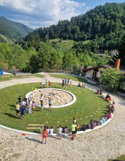 Copiii din gradinita privata se bucură de activități în aer liber în jurul unei zone circulare cu iarbă, înconjurată de copaci și munți, sub un cer parțial noros.