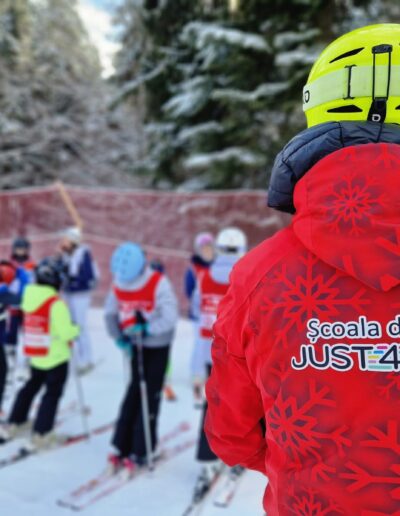 Un instructor de schi cu jachetă roșie și cască galbenă stă în fața copiilor de la o gradinita privata, învățându-i să schieze pe o pârtie înzăpezită.