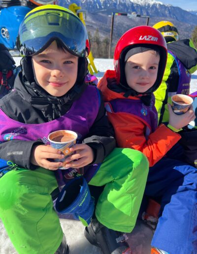 Doi copii de la o gradinita privata stau pe zăpadă în echipament de schi, savurând cești de ciocolată caldă. În spatele lor, munții înzăpeziți și cerul albastru creează un fundal de iarnă perfect.