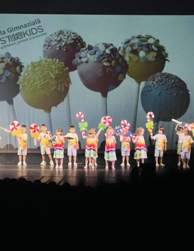 Un grup de copii de la o gradinita privata din sectorul 3, imbracati in tinute colorate, tin pe scena obiecte de recuzita din dulciuri, cu o imagine mare de prajituri si text proiectat in fundal.