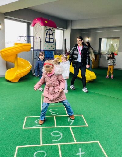 Copii și adulți se joacă pe un teren de joacă cu tobogane și un joc de șotron pe o podea verde în interiorul zonei acoperite a unei gradinita privata sector 3.