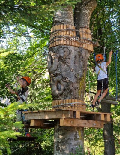 Doi copii purtând căști navighează pe un traseu de frânghii în vârful copacilor, stând pe o platformă de lemn atașată la un copac mare - o activitate aventuroasă în aer liber de care se bucură adesea copiii de la o gradinita privata din sectorul 3.