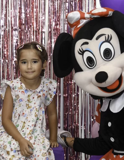 O tânără într-o rochie cu flori stă lângă o persoană într-un costum Minnie Mouse la o gradinita privata sector 3, cu baloane roz și serpentine metalice în fundal.