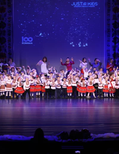 Un grup mare de copii de la o gradinita privata joacă pe scenă în costume coordonate, ghidați de supraveghetori adulți și lumini mov, creând un eveniment școlar memorabil.