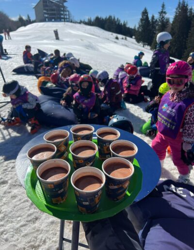 O tavă cu nouă cești de ciocolată caldă este ținută în prim-plan, în timp ce copiii de la o gradinita privata stau pe zăpadă în fundal, lângă o pârtie de schi.