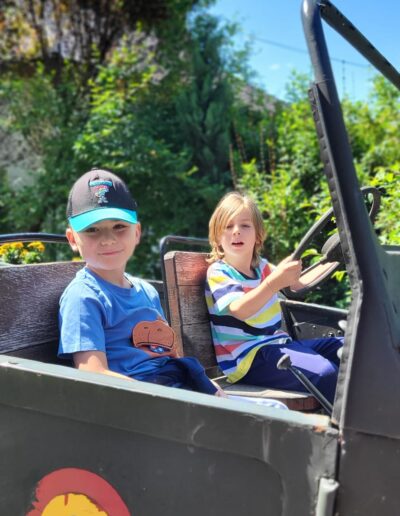 Doi copii mici stau într-un vehicul decapotabil la o gradinita privata; unul este pe scaunul șoferului, ținând volanul, în timp ce celălalt stă lângă ei. În fundal se află o verdeață luxuriantă.