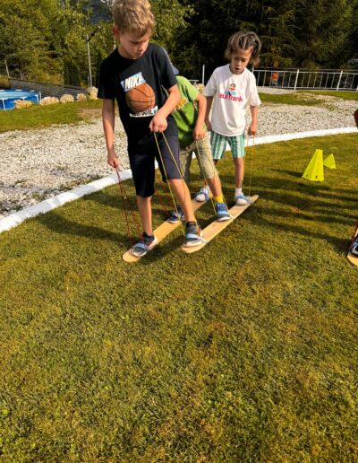 Trei copii de la o gradinita privata stau pe scânduri de lemn cu frânghii, colaborând pe un teren în aer liber cu iarbă, lângă conuri și o cărare de pietriș.