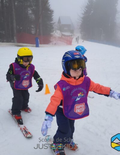 Copii tineri de la o gradinita privata sector 3, purtând căști și echipament de schi, practică schiatul pe o pârtie înzăpezită, urmând o linie de conuri portocalii. Trei sigle sunt afișate în partea de jos a imaginii.