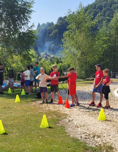 Copii și adulți se adună în aer liber într-o zi însorită, participând la o activitate organizată de tragere cu arcul - un eveniment des întâlnit la o gradinita privata sector 3 - marcată de conuri galbene pe o zonă înierbată de pe un deal împădurit.