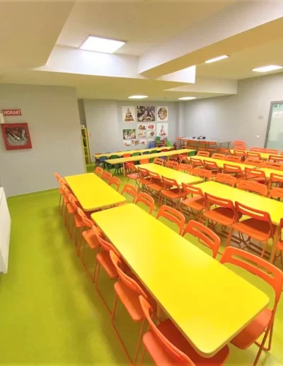 O încăpere luminată cu mese galbene și scaune portocalii dispuse în rânduri, probabil amenajată ca o cantină sau o sală de clasă într-o gradinita privata din sectorul 3. Pe pereți sunt vizibile afișe și o cutie de protecție împotriva incendiilor.